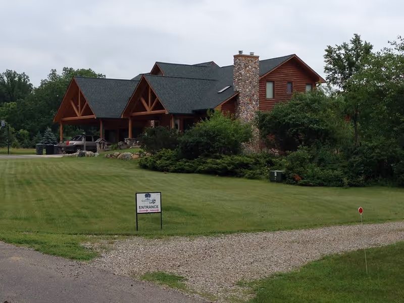 A large lodge-style building with a stone chimney and covered entrance set behind a grassy lawn and driveway.