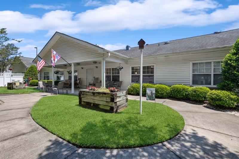 Outdoor view of a single-story senior living facility with a covered porch area featuring chairs and tables. The building is white with multiple windows, and there are two flags displayed on the porch. The foreground shows a circular grassy area with a wooden planter box filled with flowers and a birdhouse on a pole. The sky is partly cloudy and the surrounding area is landscaped with bushes and trees.