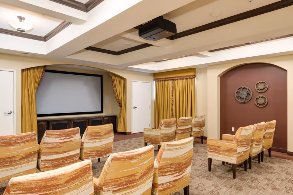 A small screening room with rows of patterned upholstered chairs facing a large projection screen framed by gold curtains.