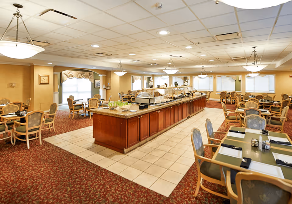 A spacious dining room in a senior living facility with multiple tables and chairs arranged around a central buffet counter. The room features warm lighting with hanging light fixtures, carpeted flooring with a patterned design, and large windows with curtains allowing natural light to enter.