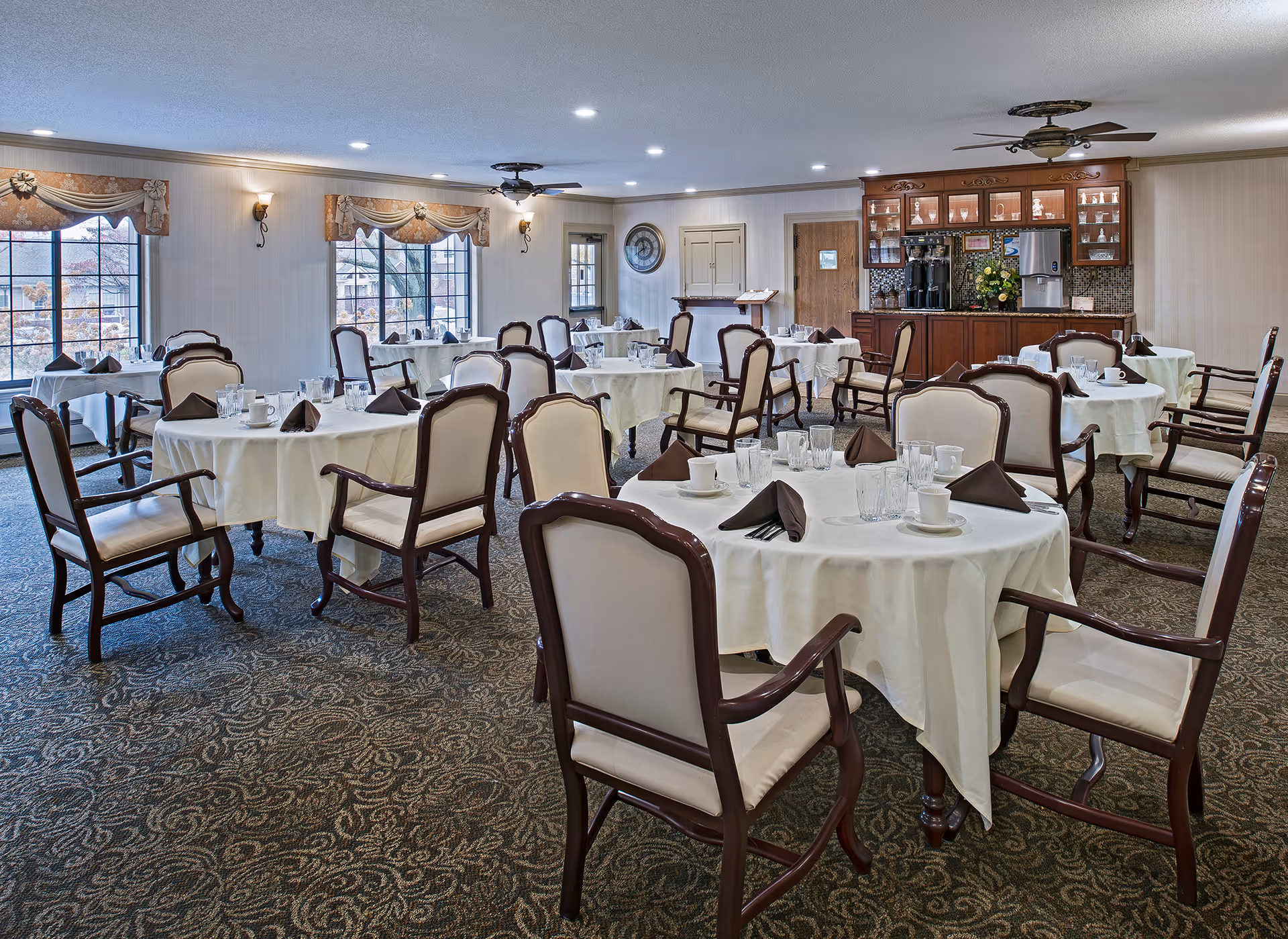 A dining room with multiple round tables covered with white tablecloths, each set with glasses, cups, and folded brown napkins. The room has large windows with decorative valances, wooden chairs with cushioned seats and backs, a carpeted floor with a patterned design, ceiling fans, and a wooden cabinet with a coffee and beverage station in the background.