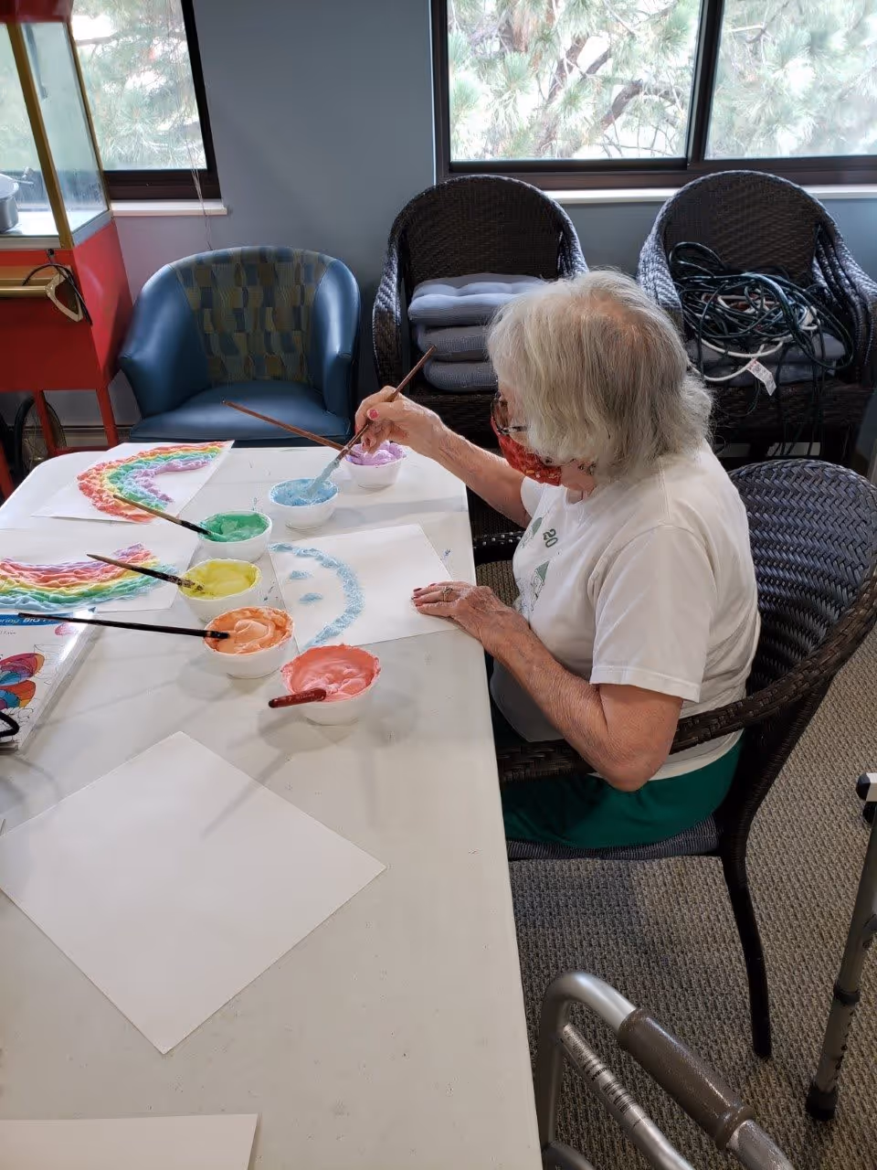 An elderly woman with white hair wearing a white shirt and green pants is sitting at a table painting with colorful paints. Several bowls of paint in different colors and paintbrushes are on the table, along with sheets of paper, one of which has a partially painted blue smiley face. Behind her are chairs and windows showing trees outside.