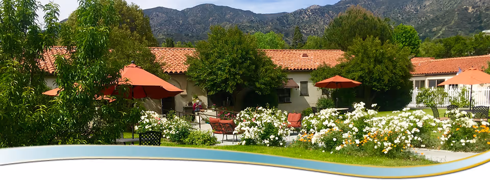 Outdoor garden area at Foothill Retirement with blooming white and yellow flowers, green trees, red patio umbrellas, and seating benches. In the background, there are buildings with red tile roofs and mountains under a blue sky.