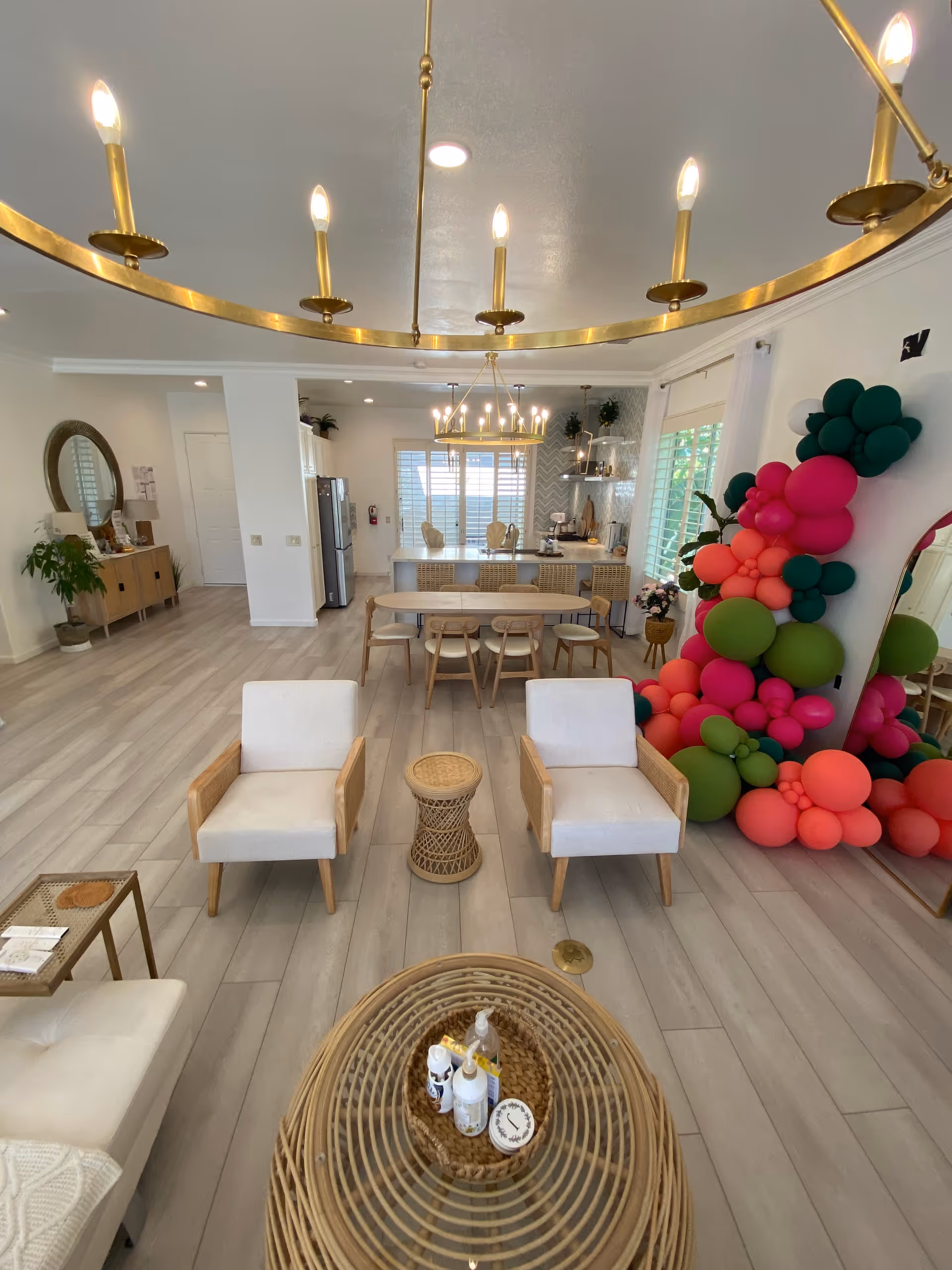 A bright and spacious living area with light wood flooring and white walls. The room features two white cushioned armchairs with wooden frames, a small round wicker side table between them, and a wicker coffee table with various bottles and containers on top. In the background, there is a dining table with chairs and a kitchen area with a refrigerator and shelves. A large mirror decorated with colorful balloons in shades of pink, green, and red is positioned on the right side. Two modern gold chandeliers hang from the ceiling.
