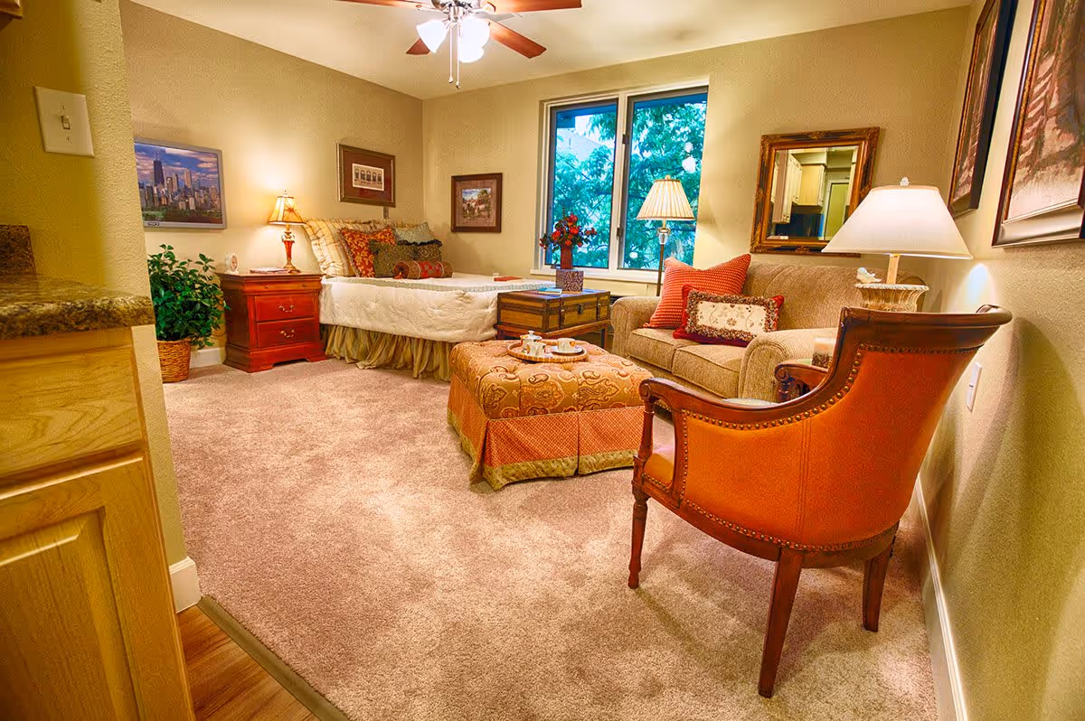 A cozy room in a retirement community featuring a single bed with multiple pillows, a wooden nightstand with a lamp, a beige sofa with decorative pillows, an upholstered ottoman with a tea set on top, a wooden armchair with orange upholstery, a window showing green trees outside, and warm lighting from ceiling fan lights and lamps.