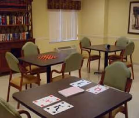 A room with several tables and chairs arranged for card games. Playing cards and a deck are placed on the nearest table. There is a bookshelf filled with books against one wall and a window with blinds and a valance. The room has a calm, neutral color scheme.