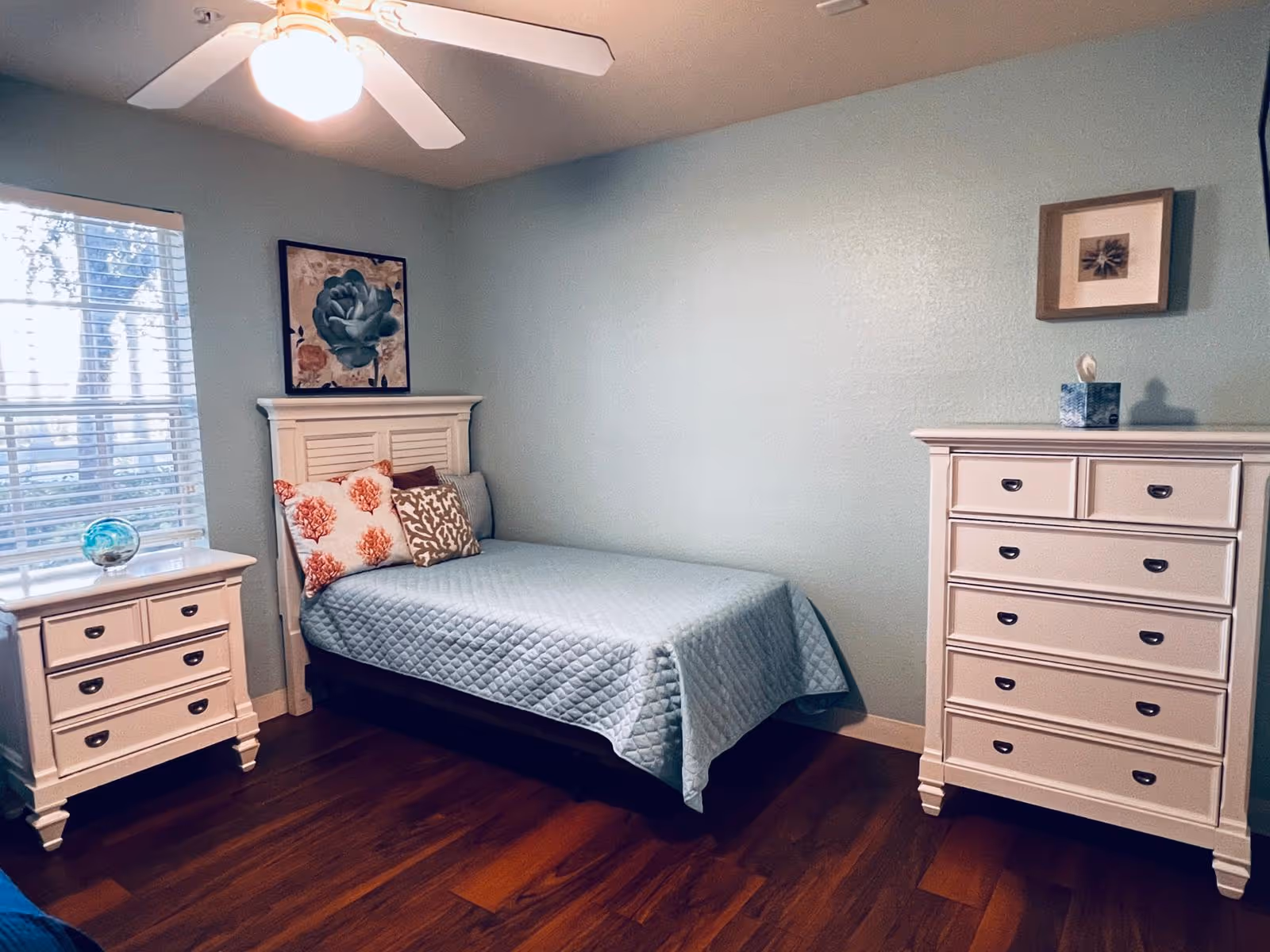 A bedroom with light blue walls and wooden flooring featuring a single bed with a light blue quilt and decorative pillows. There is a white nightstand with drawers and a decorative glass orb near a window with blinds on the left side. On the right side, there is a white chest of drawers with a tissue box on top and a framed picture hanging above it. A ceiling fan with a light is visible on the ceiling.