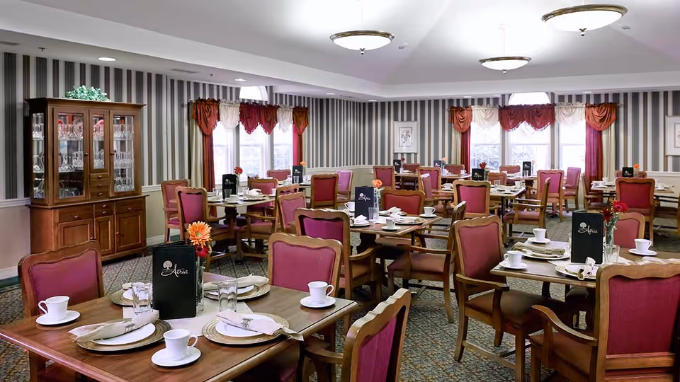 A dining room with multiple wooden tables and chairs featuring red upholstery. Each table is set with white plates, cups, glasses, and napkins, along with a small flower vase and a menu. The room has striped wallpaper, large windows with red and cream curtains, and a wooden cabinet displaying glassware.