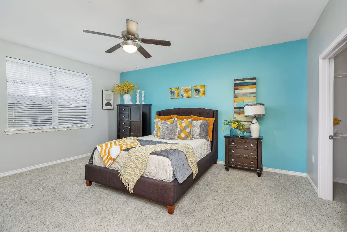 A bright and cozy bedroom with a dark upholstered bed adorned with multiple yellow, gray, and white pillows and blankets. The room features a light blue accent wall behind the bed, a ceiling fan with lights, a window with white blinds, a dark wooden dresser with decorative vases and flowers, and a matching nightstand with a lamp and floral arrangement. The floor is carpeted in a light gray color.