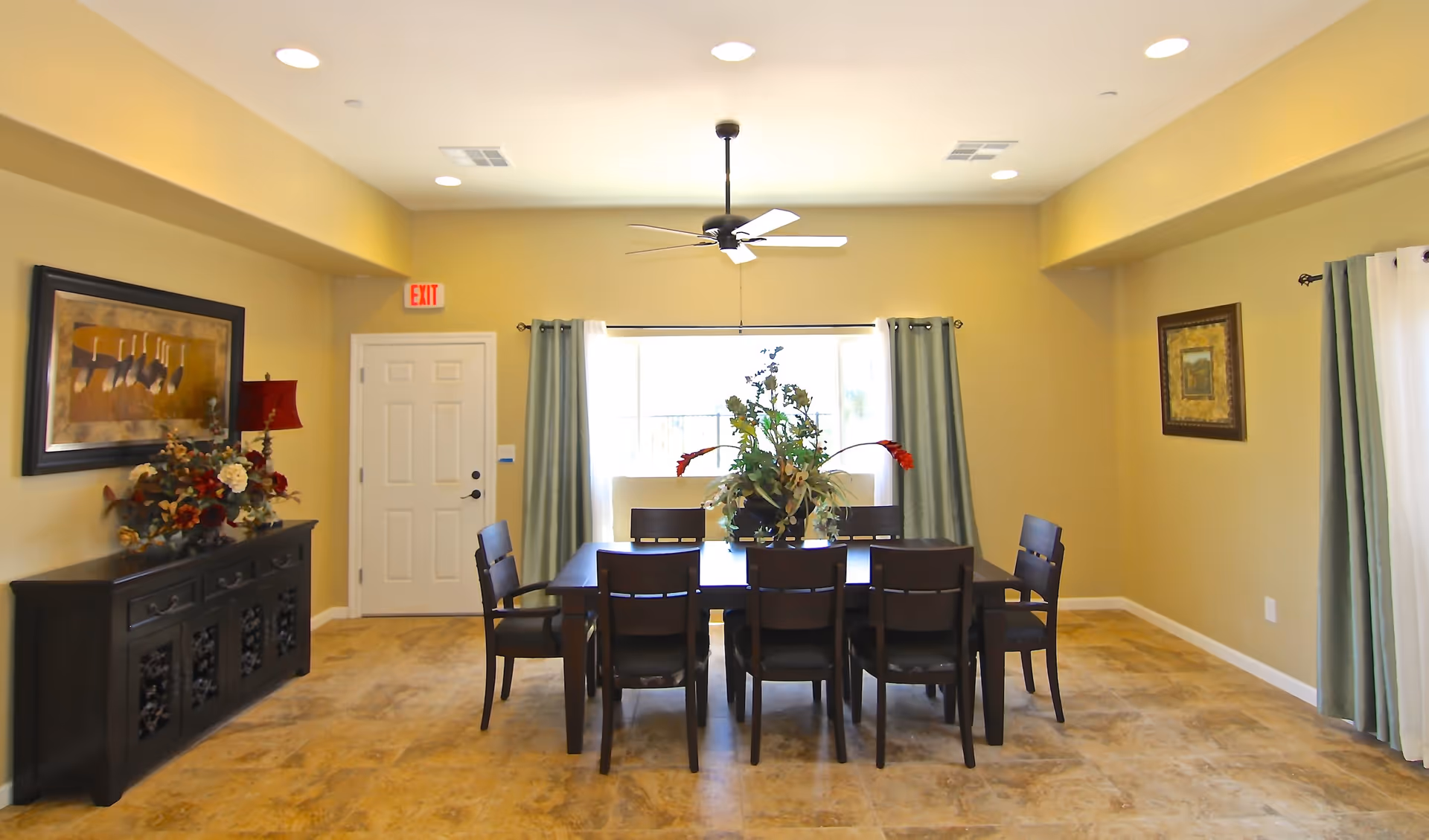 A dining room with a dark wooden table surrounded by eight matching chairs. A large floral arrangement sits in the center of the table. The room has beige walls, a ceiling fan with lights, and a window with light green curtains letting in natural light. There is a dark wooden sideboard with a lamp and floral decoration on the left wall, and framed artwork on both side walls. The floor is tiled with a light brown pattern.