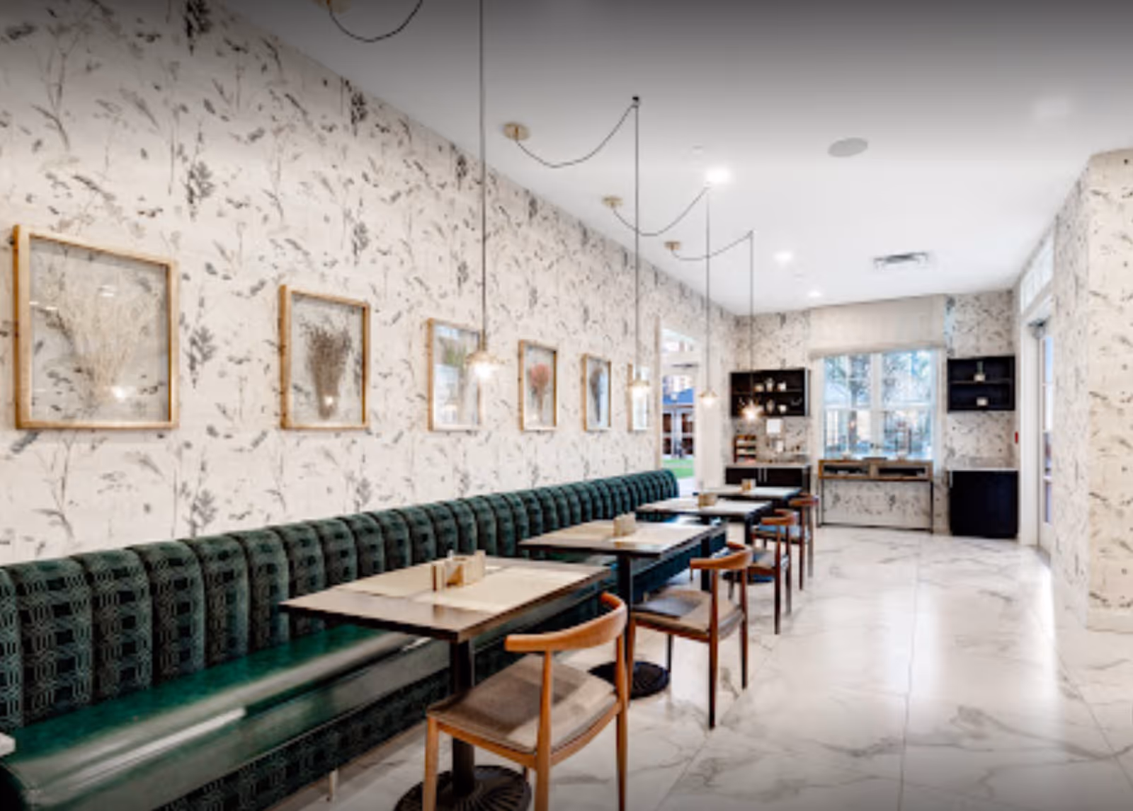 A bright dining area with a long green upholstered bench along one wall, several wooden tables and chairs arranged in a row, floral patterned wallpaper, framed botanical artwork on the wall, pendant lights hanging from the ceiling, and large windows letting in natural light.