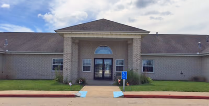 Single-story brick healthcare building front with a covered entrance, double glass doors, small lawn, and a handicapped parking sign.