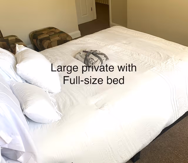 Full-size bed with white bedding and pillows in a private bedroom with a doorway and ottoman visible.