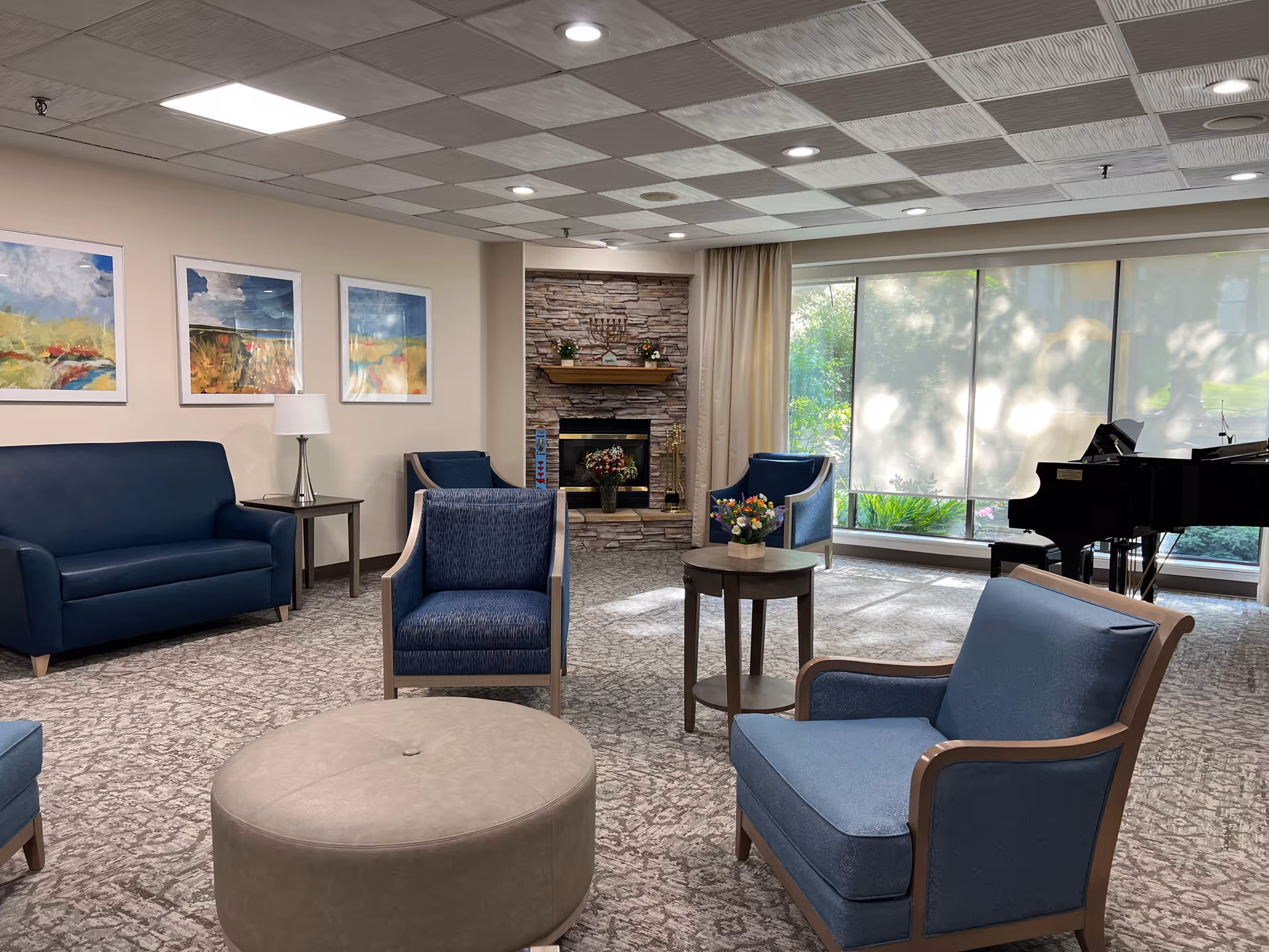 A cozy living room area with blue upholstered chairs and a sofa arranged around a round beige ottoman and a small wooden table with a flower arrangement. The room features a stone fireplace with decorative items on the mantel, three colorful landscape paintings on the wall, large windows with shades partially drawn, and a black grand piano near the windows. The ceiling has a checkered pattern with recessed lighting.