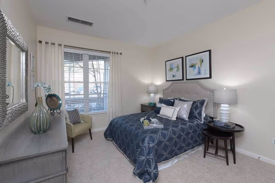 A bedroom with a bed covered in a blue patterned comforter and multiple pillows. There are two framed abstract paintings above the bed's upholstered headboard. On either side of the bed are nightstands with white lamps. A window with white curtains and blinds is on the left wall, next to a beige armchair. A dresser with decorative items and a large mirror is also visible.