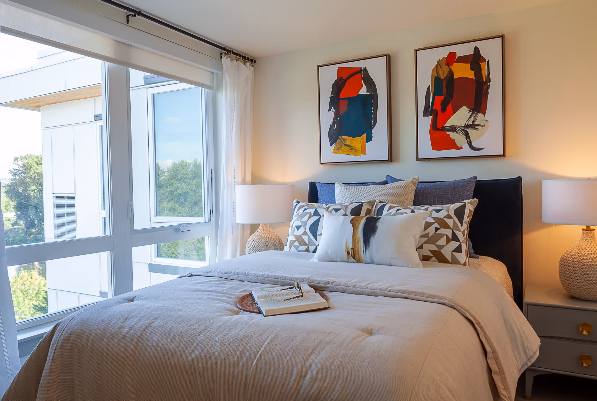 A neatly made bed with multiple decorative pillows and a beige comforter in a bright bedroom. Two abstract art paintings hang on the wall above the bed. There are two bedside tables with matching lamps on either side of the bed. Large windows with white curtains allow natural light to fill the room, showing a view of trees and a neighboring building outside.