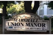 Wooden entrance sign reading "Marshall Union Manor" set among trees with a building visible behind it.