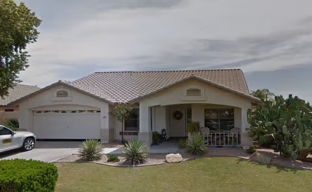 Single-story house with a tiled roof, a two-car garage, a small front porch with seating, desert landscaping including cacti and small shrubs, and a car parked in the driveway under a cloudy sky.
