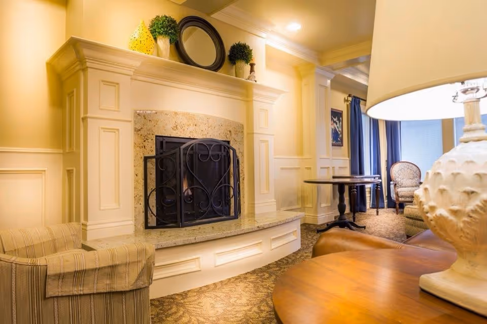 A cozy living room area with a decorative fireplace featuring a black metal screen. On the mantle above the fireplace are two small green plants, a yellow decorative object, and a round black-framed mirror. The room has warm yellow walls with white paneling, a patterned carpet, a striped armchair, a wooden table with a large white lamp, and a cushioned chair near a window with blue curtains.