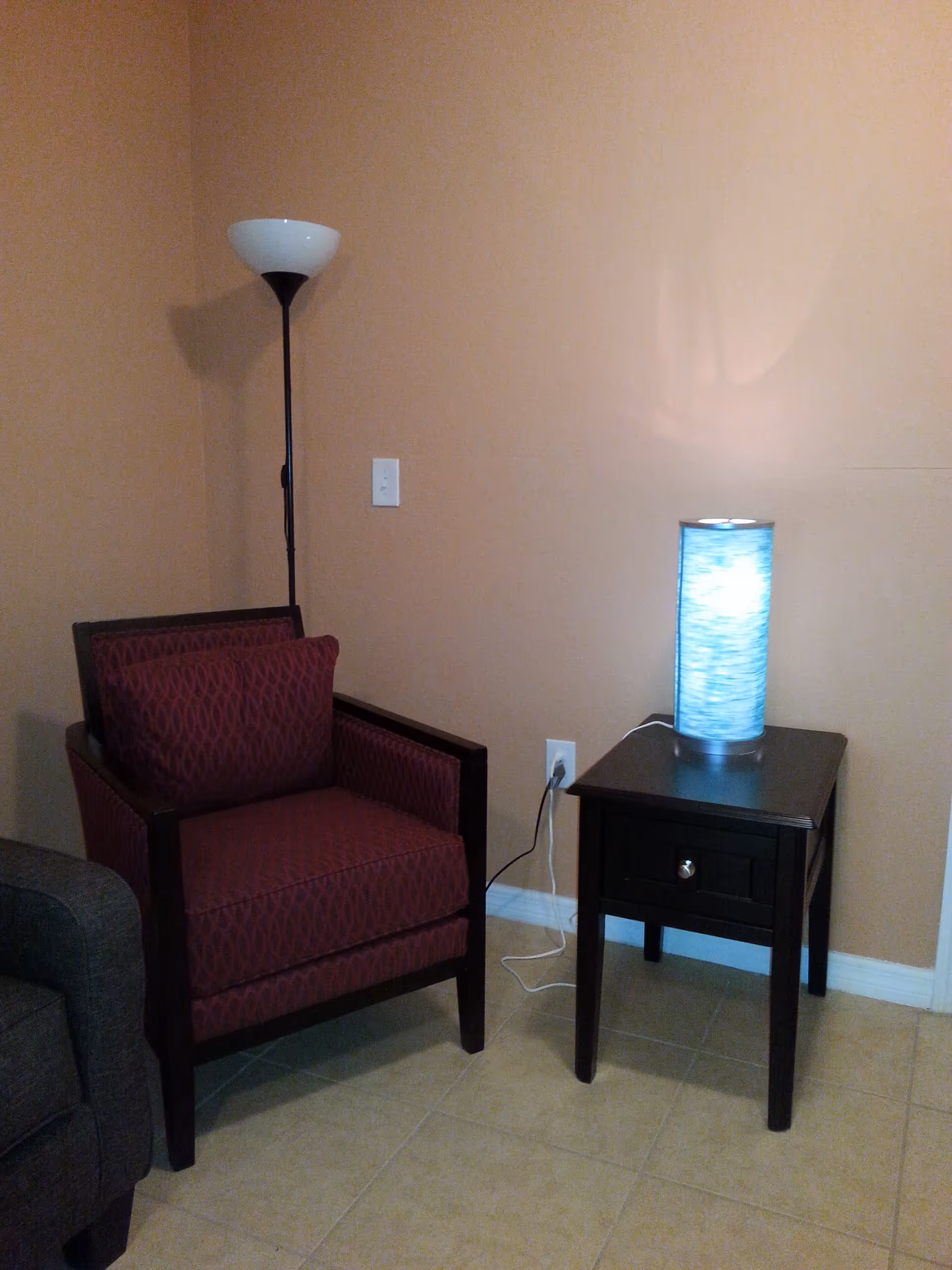 A cozy corner of a room with a maroon patterned armchair, a black wooden side table with a drawer, and a cylindrical blue lamp on the table. A tall floor lamp with a white shade stands behind the armchair against a beige wall.