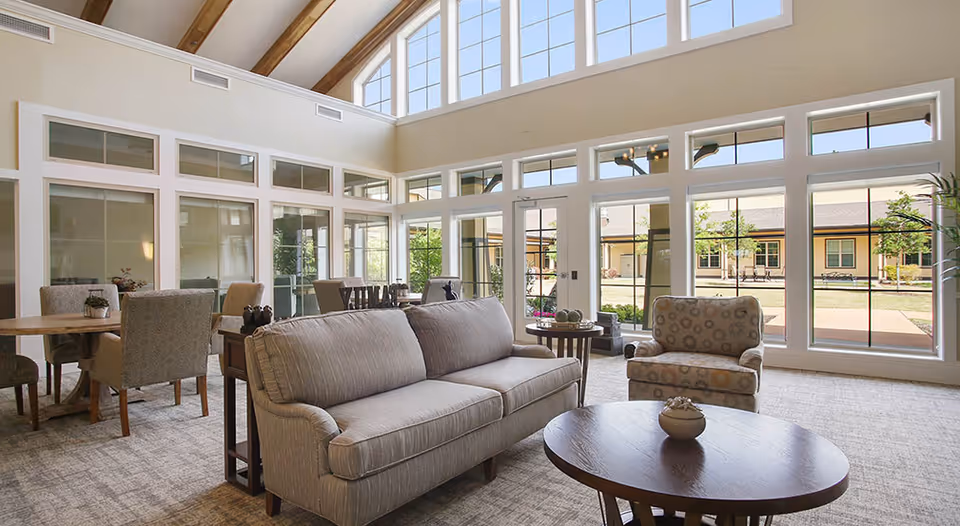 A bright and spacious living room area with large windows allowing natural light to fill the space. The room features a beige sofa, a patterned armchair, a round wooden coffee table with a decorative bowl, and a dining table with upholstered chairs in the background. The ceiling has exposed wooden beams and there is a view of an outdoor courtyard through the windows.
