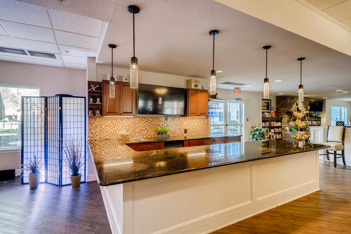 A modern kitchen area in a senior living facility with a long granite countertop island, pendant lights hanging from the ceiling, wooden cabinets, a mosaic tile backsplash, and a flat-screen TV mounted on the wall. There are decorative plants and a tiered fruit basket on the counter. In the background, there is a bookshelf, a stone fireplace, and seating areas with chairs.