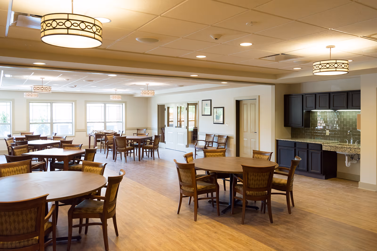 Spacious dining room with round wooden tables and chairs, large windows, and a kitchenette along one wall.