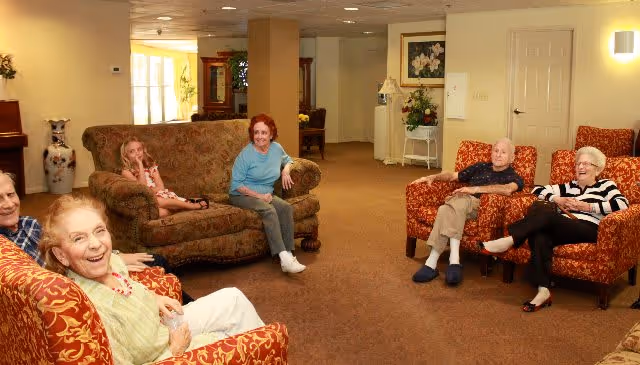 A group of elderly people and a young girl sitting and smiling in a cozy living room area with patterned armchairs and a sofa. The room has warm lighting, carpeted floor, and decorative elements including a large vase and framed artwork on the walls.