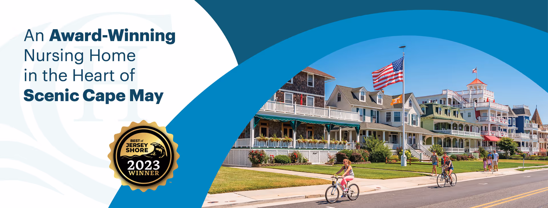 A scenic street view of a coastal town with large, colorful Victorian-style houses and an American flag flying on a flagpole. People are riding bicycles and walking along the sidewalk on a sunny day.