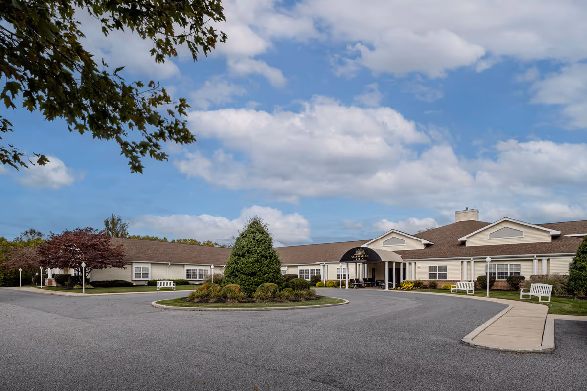 Exterior view of Celebration Villa of York, a single-story building with a circular driveway and landscaped greenery including bushes and trees under a partly cloudy sky.
