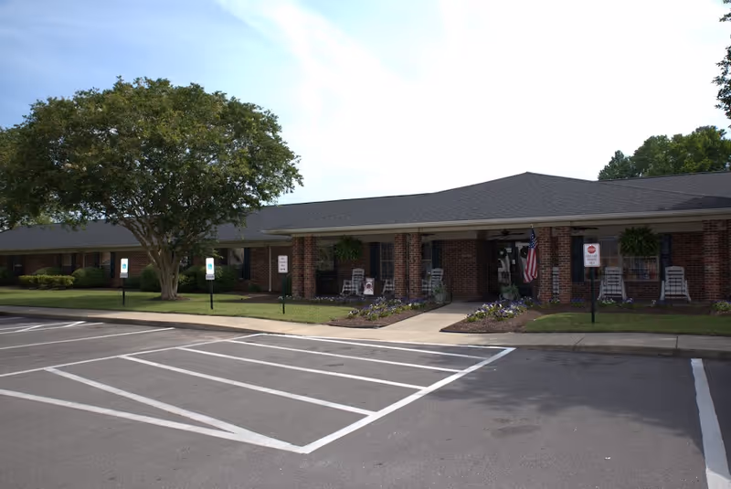 Single-story brick senior living facility front with a walkway, parking spaces, landscaping, and an American flag.