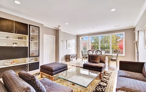 A bright and spacious living room with large windows letting in natural light. The room features a glass coffee table on a patterned rug, surrounded by dark upholstered chairs and a matching ottoman. In the background, there is a dining area with a wooden table and chairs near the windows, and built-in shelving on the left wall displaying decorative items.