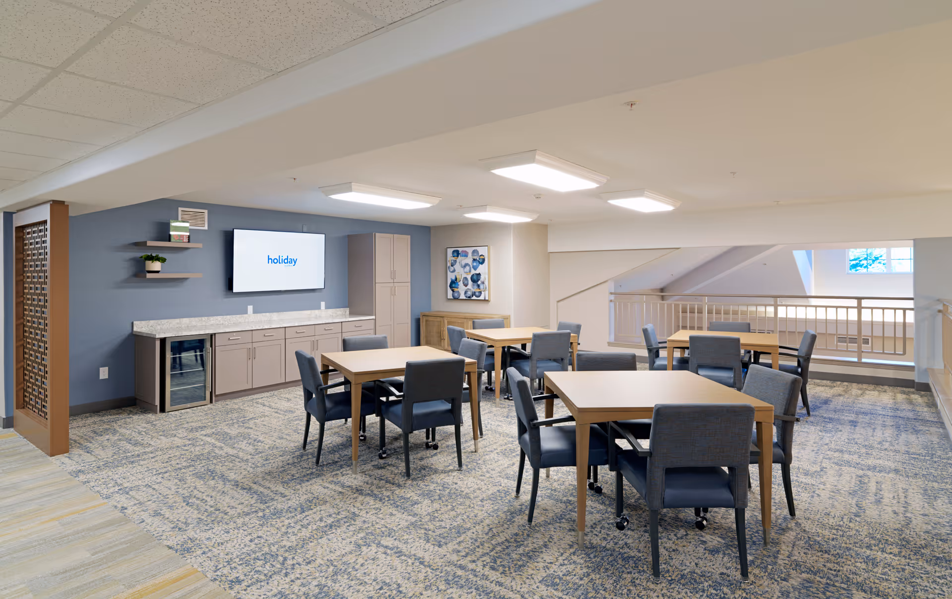 A well-lit common area with several wooden tables and blue upholstered chairs on a patterned carpet. There is a blue accent wall with a mounted flat-screen TV displaying the word 'holiday', shelves with decorative items, and cabinets with a countertop. The space has a modern ceiling with recessed lighting and a railing overlooking a lower level.