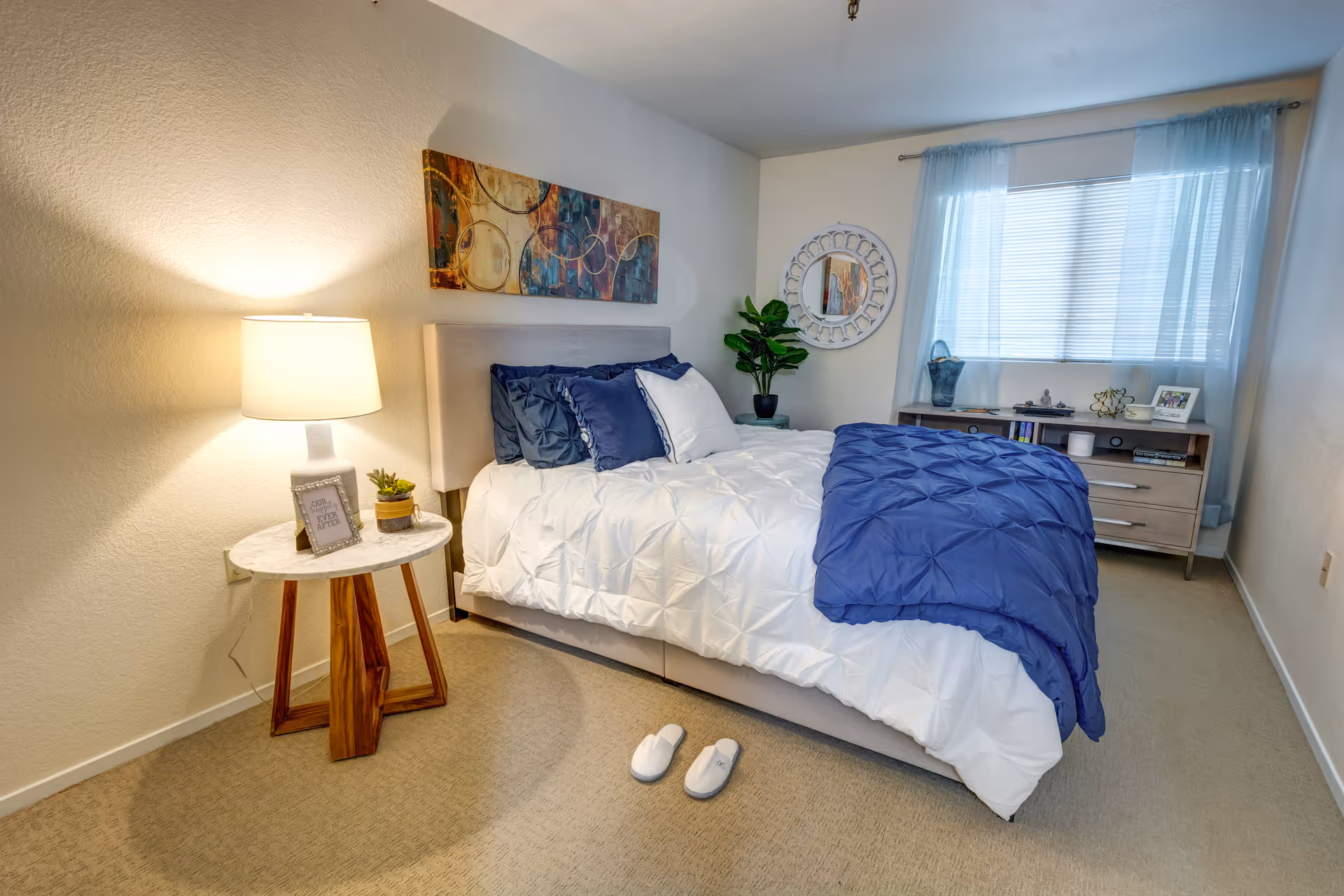 A cozy bedroom with a neatly made bed featuring white and blue bedding, a pair of white slippers on the carpeted floor, a wooden side table with a lamp and small decor items, a decorative round mirror on the wall, a potted plant, and a dresser under a window with sheer blue curtains.