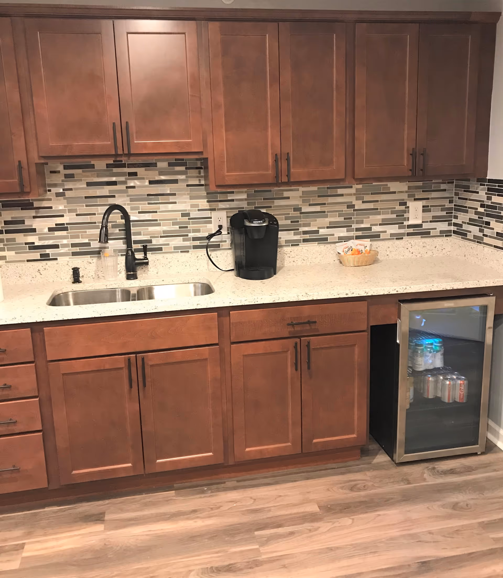 A kitchen area with wooden cabinets, a double stainless steel sink with a black faucet, a Keurig coffee maker, a small basket with coffee supplies, and a mini refrigerator stocked with beverages. The backsplash features a mosaic tile design in shades of gray, white, and black, and the floor is wood-patterned.