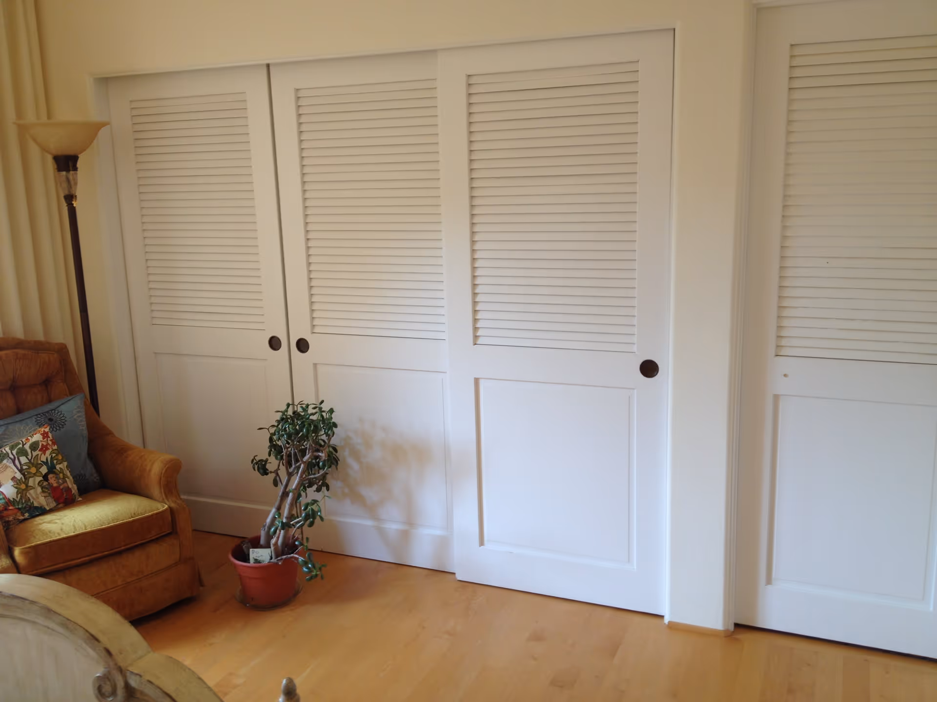 White louvered sliding closet doors in a room with a brown armchair, floor lamp, and a potted plant on a wood floor.