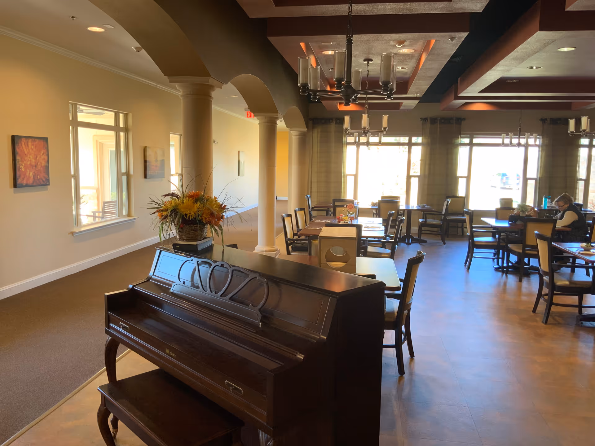 Spacious senior living dining area with a piano in the foreground, tables and chairs, columns, and large windows.