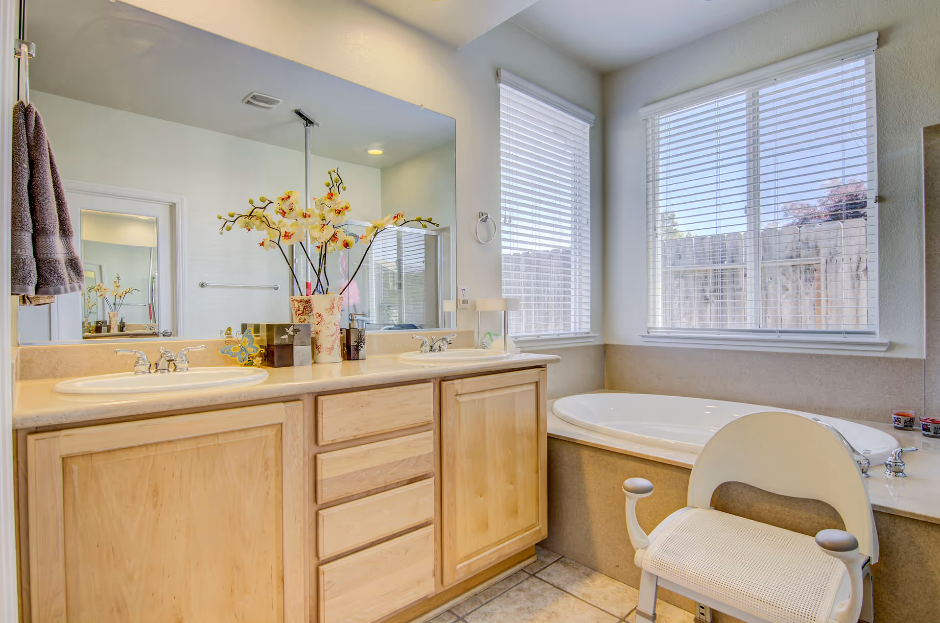 A bright bathroom featuring a double sink vanity with wooden cabinets and a large mirror above. There is a decorative vase with yellow orchids on the countertop. Next to the vanity is a large bathtub positioned under two windows with white blinds. A white shower chair is placed in front of the bathtub. The floor is tiled and the walls are painted light beige.