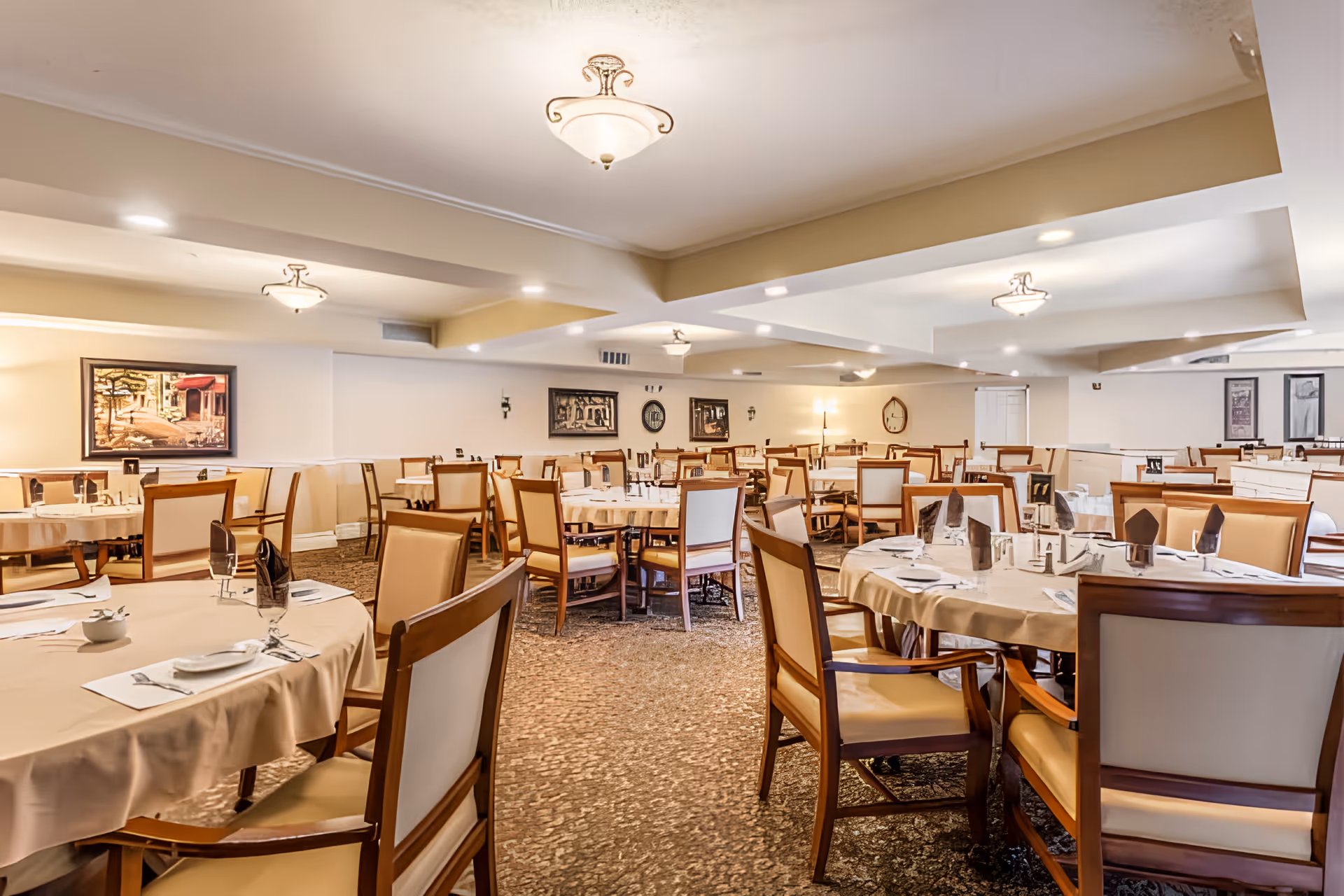 A spacious dining room with multiple round tables covered with beige tablecloths, each set with plates, napkins, and silverware. The room has beige walls adorned with framed artwork and is illuminated by ceiling lights. Wooden chairs with beige cushions surround the tables.