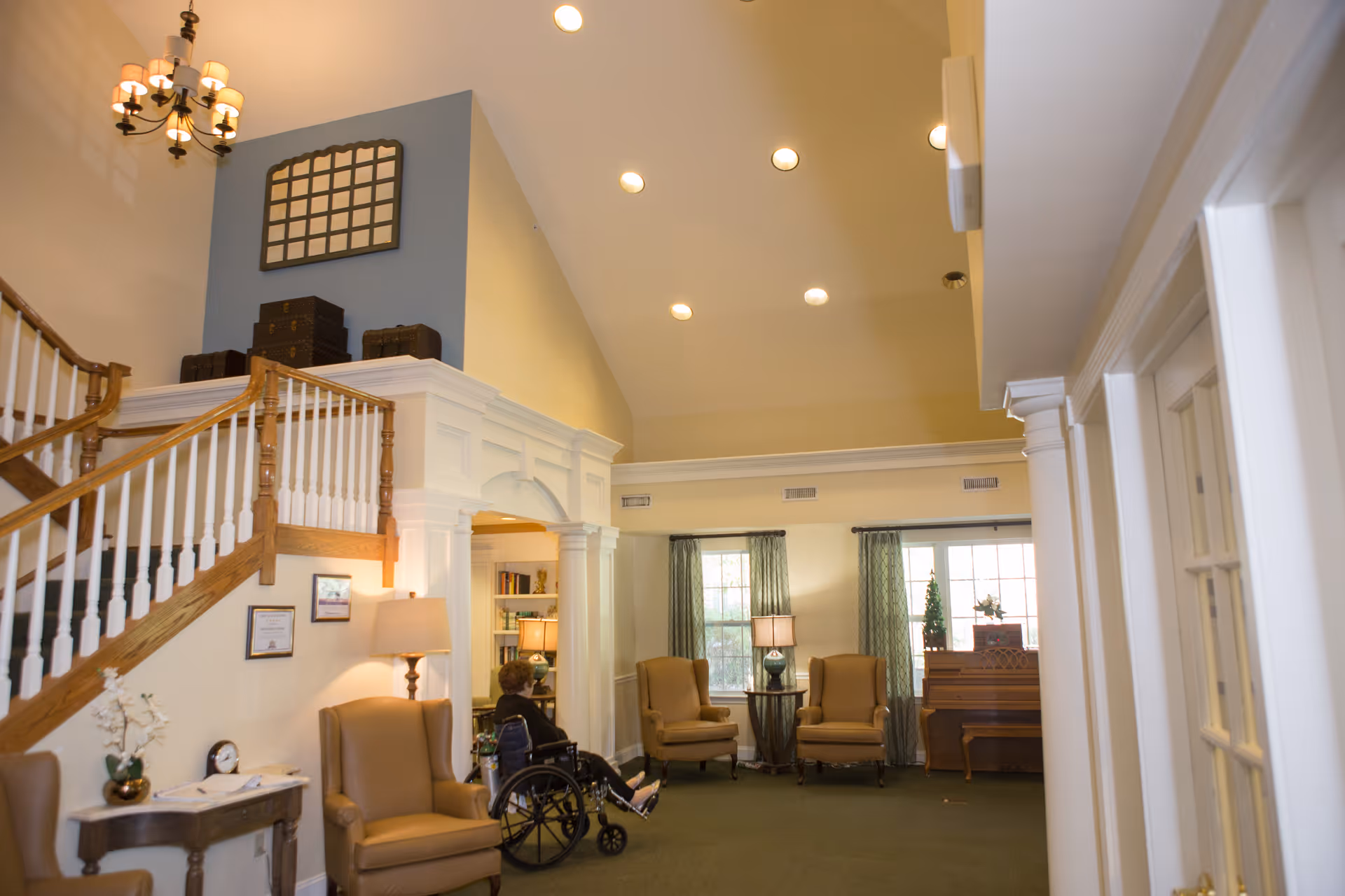 Sunlit senior living common room with high ceilings, a staircase, seating, a piano and a resident in a wheelchair.