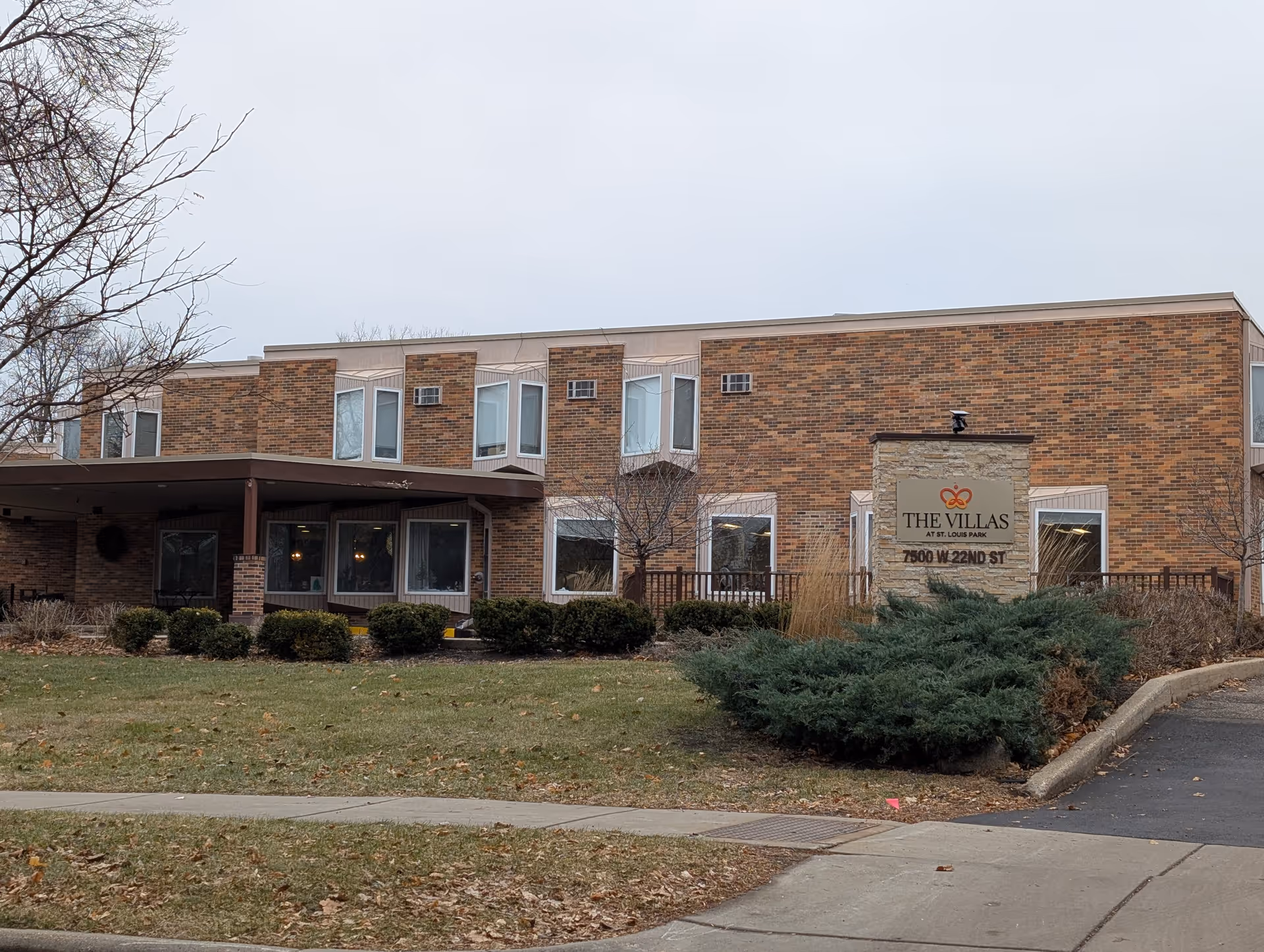 Exterior view of The Villas at St. Louis Park building, a two-story brick structure with multiple windows and a covered entrance. There is a sign in front displaying the facility's name and address, surrounded by shrubs and a lawn with a sidewalk and driveway nearby.