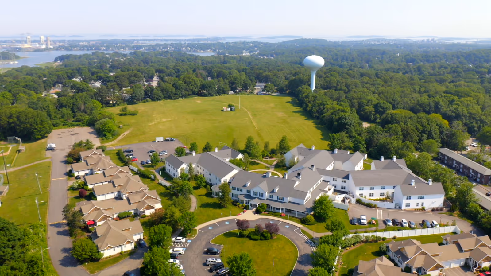 Aerial view of Allerton House Weymouth senior living facility showing multiple buildings surrounded by trees and greenery, a large open field, a circular driveway with parked cars, and a water tower in the background.