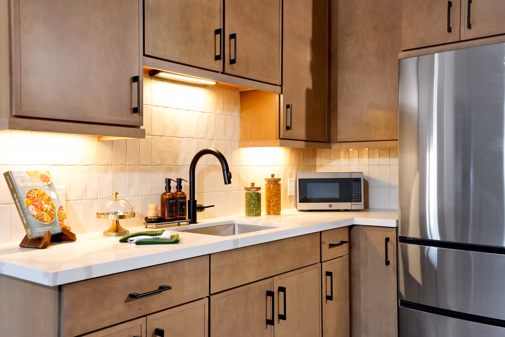 Modern kitchen countertop with wood cabinets, undermount sink and black faucet, microwave, and stainless steel refrigerator.