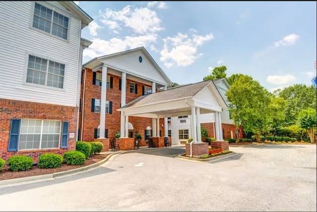 Exterior view of a senior living facility named Carolina Gardens at Kathwood, featuring a brick and white-paneled building with large windows, a covered entrance supported by white columns, and surrounding greenery under a partly cloudy sky.