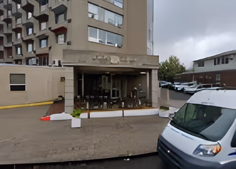 Exterior view of a multi-story building with a covered entrance area featuring glass doors and some outdoor seating. A white van is parked in the foreground on the right side. Other buildings and parked cars are visible in the background under a cloudy sky.