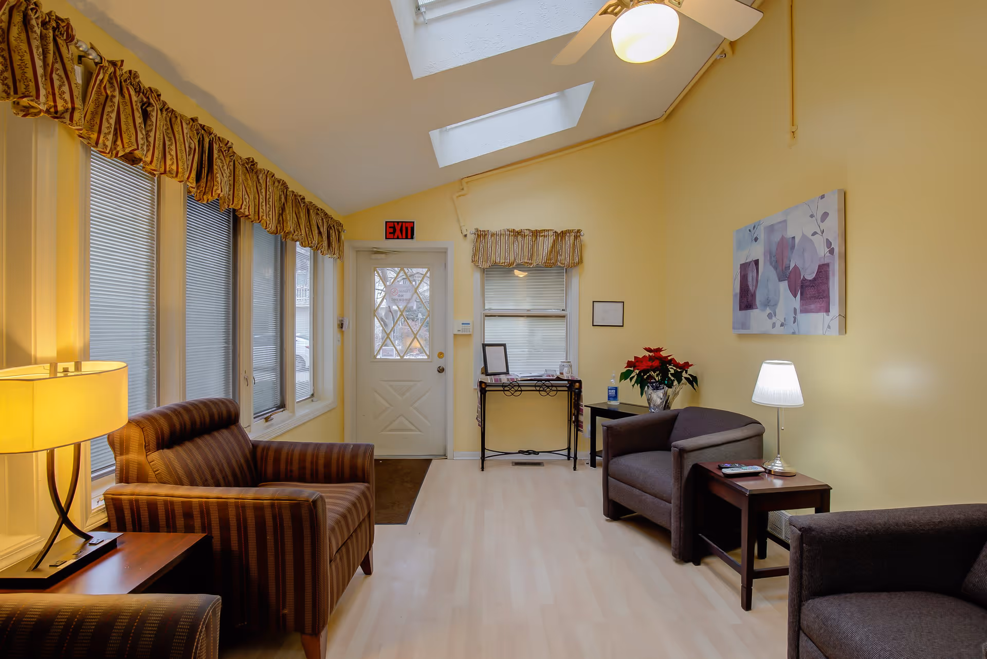 Cozy, well-lit common room with striped and upholstered armchairs, side tables and lamps, skylights, and an exit door.