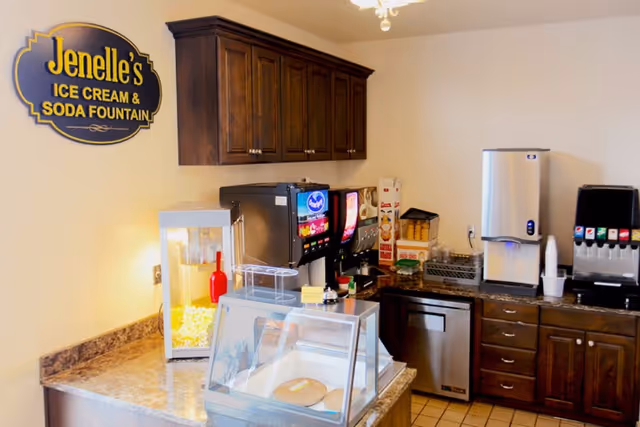 A kitchenette-style snack bar with a 'Jenelle's Ice Cream & Soda Fountain' sign, popcorn machine, beverage dispensers, and a pastry display case.