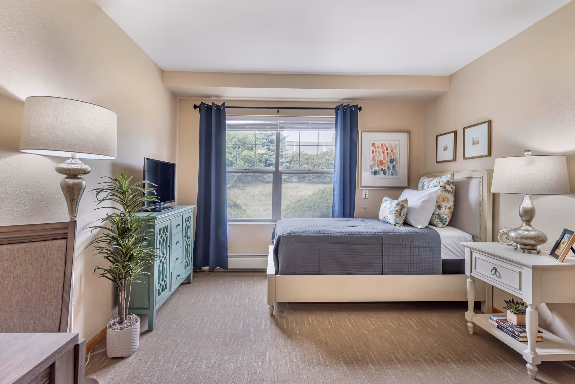 A neatly arranged bedroom with a bed featuring gray bedding and floral pillows, two bedside tables each with a lamp, a green cabinet with a TV on top, a potted plant, and a large window with blue curtains showing greenery outside.