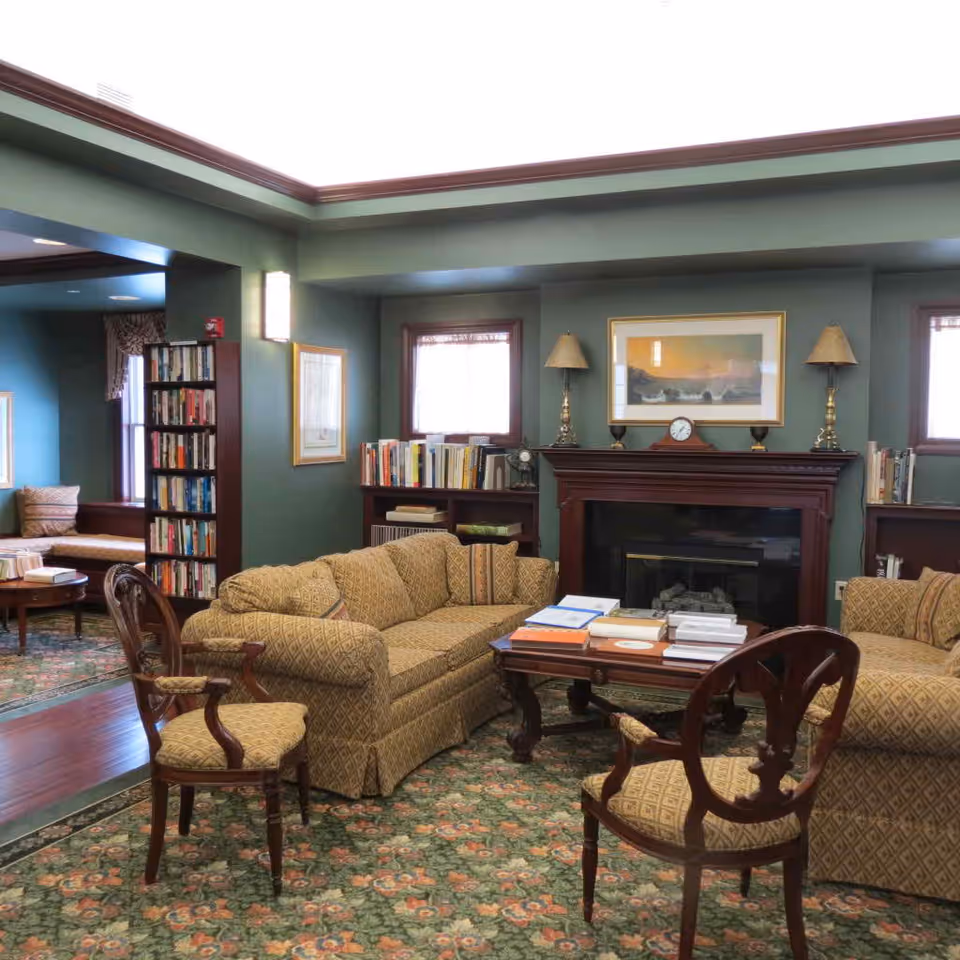 A cozy living room area in Meadow Ridge with patterned upholstered sofas and chairs arranged around a wooden coffee table stacked with books. The room features a fireplace with a clock and lamps on the mantel, bookshelves filled with books, green walls, and a floral carpet. There is a window with curtains and framed artwork above the fireplace.