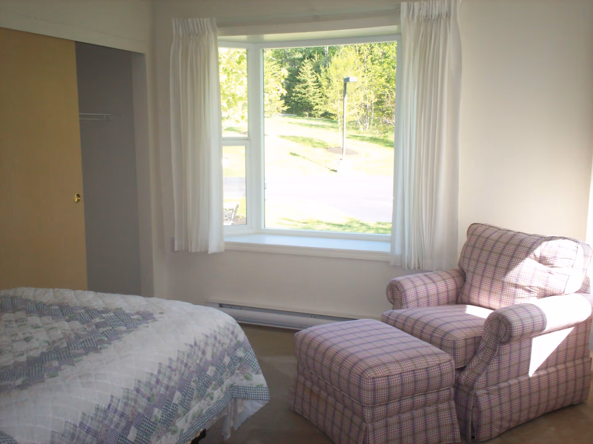 Sunlit bedroom with a large window, a plaid armchair and ottoman next to a bed.